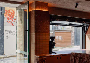 Interior view of a modern industrial-style coffee shop featuring a corten steel pillar, terracotta tile wainscoting, a black coffee grinder on a stone countertop, and a large glass door opening to an urban alley with graffiti art.
