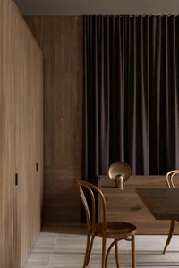 Modern minimalist dining area featuring warm walnut wood cabinetry, a bentwood chair, a wooden sideboard with a brass table lamp, and dark brown curtains.
