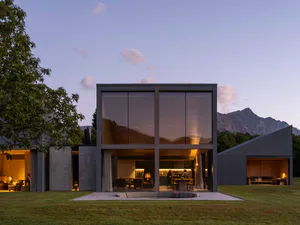 Modern glass house with large transparent windows reflecting mountains, open plan interior with people in kitchen and living area, outdoor concrete patio with curved sofa on green lawn at dusk.
