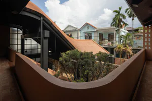 View from a curved terracotta rooftop terrace with lush greenery overlooking neighboring houses and palm trees under a blue sky.