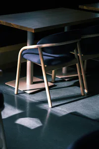 Modern ash wood dining chair with curved backrest and navy blue fabric seat and back, positioned under a solid wood table with dramatic directional lighting casting strong shadows on a polished concrete floor.
