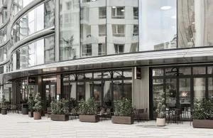 Modern building with curved glass facade and outdoor restaurant seating area featuring potted plants and metal furniture on a paved plaza.