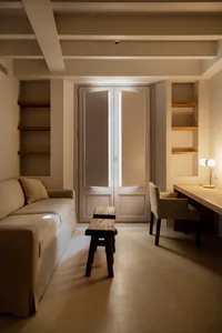 A minimalist living room with a light beige sofa, two rustic wooden benches in the center, built-in wooden shelves on both sides, a wooden desk with a fabric chair and modern lamp on the right, and double doors leading to a balcony in the background.