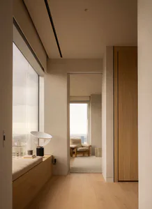 Modern minimalist hallway with light oak wood flooring, built-in window bench with cushion, contemporary table lamp, and a view of the city through large windows. A wooden door is visible on the right and a seating area appears in the background room.