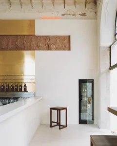 Interior view of a minimalist bar with white walls, a brass panel, terracotta classical frieze, orange neon sign reading SOCIALE, wooden stool, and glass-door refrigerator.