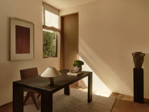 Minimalist home office corner featuring a dark walnut desk, upholstered chair, table lamp, potted plant, framed artwork, and an ancient-style vase on a black pedestal, bathed in warm natural light.