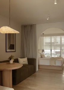 Modern minimalist interior featuring a living area with brown modular sofa, round oak table, and large arched opening revealing a bedroom with platform bed and large windows.