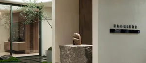 Modern minimalist entryway featuring a large natural stone pedestal with a wooden sculpture, glass door revealing a wooden display cabinet, and indoor greenery.