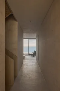 Long minimalist concrete hallway with large floor-to-ceiling windows revealing an ocean view and a green lounge chair at the end.