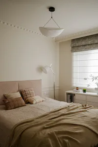 Cozy neutral bedroom with a large textured pinkish headboard, layered beige and brown pillows, a mustard linen throw, sculptural white pendant light, and a white console table under a window with roman blinds.