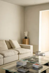 A minimalist living room featuring a cream-colored sectional sofa, a transparent glass coffee table with stacks of design books, a wooden pedestal displaying a white stone torso sculpture, and a light wood doorway in the background.