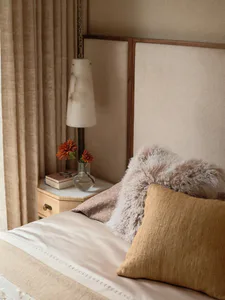 Close-up of a serene bedroom corner featuring a upholstered beige headboard, mustard corduroy pillow, fluffy alpaca wool cushion, marble pendant lamp, and a wooden nightstand with marble top holding a glass vase with orange flowers.