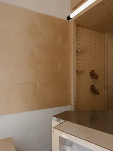 Modern minimalist entryway corner with light plywood wall paneling, a wooden pegboard holding two pairs of brown shoes, a dark countertop, and a long cylindrical pendant light.