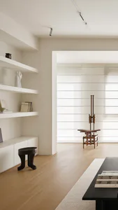 Minimalist living room interior with light oak wood flooring, white built-in floating shelves displaying books and sculptures, a dark wooden three-legged stool, and a tall wooden easel-like sculpture near a large window with horizontal blinds.
