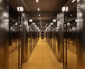 Long symmetrical corridor lined with large cylindrical stainless steel pillars and full-height mirrors creating infinite reflections on a warm golden carpet.