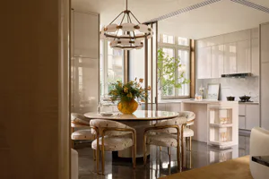 Modern luxury dining room with a round marble table, four upholstered dining chairs with gold frames, a large sculptural chandelier, and a marble kitchen island in the background.