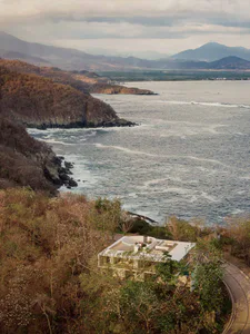 Modern concrete villa built into a forested cliffside overlooking the Pacific Ocean with distant mountains and coastline visible at sunset.
