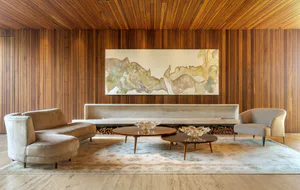 Mid-century modern living room featuring walnut wood paneling, a long travertine floating hearth with stacked firewood, two curved beige sofas, round walnut coffee tables, and a large abstract painting above the fireplace.