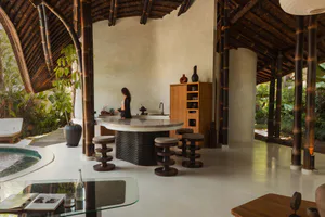 Open-air tropical pavilion with dark bamboo columns, a curved marble kitchen island, wooden bar stools, and a small pool visible on the left.