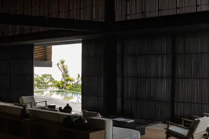 Modern tropical living room interior with dark vertical wood slat walls, light neutral sofas, wooden armchairs, black ceramic vases, and an open view to an infinity pool surrounded by tropical plants.
