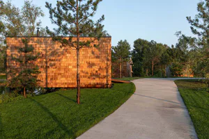 Modern cedar shingle cabin exterior with a curved concrete driveway, surrounded by pine trees and green lawn at sunset.