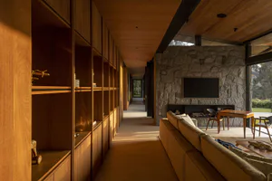 Modern living room interior featuring a long wooden hallway with built-in shelving, a large stone fireplace with mounted TV, and a beige sectional sofa in warm afternoon light.