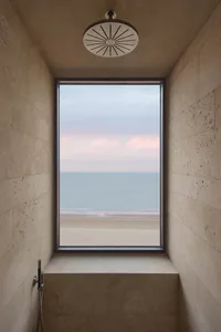 Modern minimalist shower room with large floor-to-ceiling window overlooking a sandy beach and calm ocean at dusk, featuring travertine stone walls, a large circular rain shower head, and a built-in stone bench.