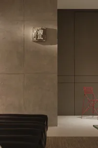 Modern minimalist interior featuring a large textured concrete wall with a stone plaque bearing the Chinese character for 'Li' or 'Propriety', a bright red wireframe bar stool, and a dark leather bench in the foreground.