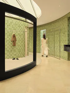 Modern boutique interior featuring curved black architectural frame, sage green ceramic tile walls, minimalist clothing display, and a blurred figure walking through the space.