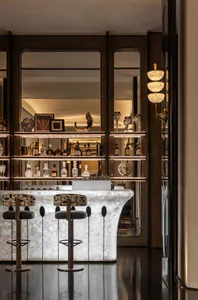 Luxurious modern home bar with a white and grey veined Calacatta marble curved counter, two patterned bar stools with dark green velvet seats, backlit dark metal and glass display shelves filled with liquor bottles and crystal glassware, and a sculptural alabaster wall sconce on the right.