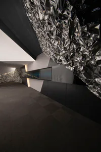 Modern art gallery interior featuring a large sculptural installation of crumpled reflective metal suspended from the ceiling, dark tiled floor, and digital display screens along the wall.