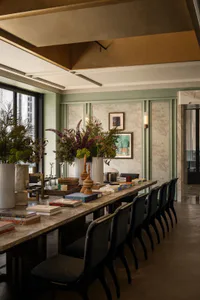 Long marble dining table with dark upholstered chairs in a sage green dining room featuring large floral arrangements in white vases, framed art, and warm ceiling lighting.