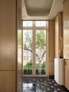 Modern luxury entryway with beige paneled walls, black marble checkered floor, tall glass doors overlooking a garden with trees, and a wooden sculpture on a cylindrical travertine pedestal.