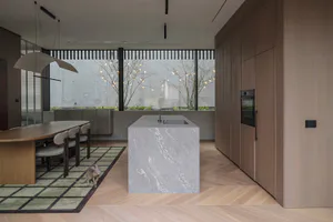 Modern minimalist kitchen with a large grey marble island, built-in oven, wooden cabinetry, a wooden dining table with chairs on a green rug, and a cat walking nearby, with large windows overlooking a garden with string lights.