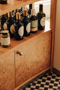 Close-up of a burl wood kitchen pantry cabinet with round knob, displaying numerous dark glass bottles of olive oil and a jar of olives on the wooden countertop.