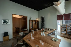 Modern Japanese restaurant interior with large L-shaped wooden dining counter, woven chairs, shoji-style sliding doors, and minimalist table settings with ceramic cups and chopsticks.