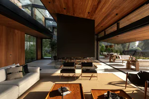 Modern open-plan living room with high wood-paneled ceiling, large black fireplace, light sofas, leather chairs, and views to outdoor dining and garden.
