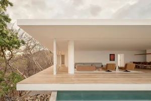 Modern open-plan living room with concrete overhang, large glass walls, mustard swivel chairs, and a sweeping view of a dry hillside and infinity pool.