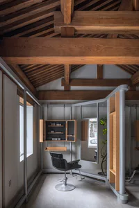 Modern industrial hair salon interior with exposed wooden ceiling beams, black leather salon chair, floating wooden cabinetry, and concrete floor.