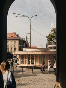 View through a stone arch of a modern curved wooden kiosk named Cheboteka on a sunny cobblestone square in Prague, with pedestrians walking by.