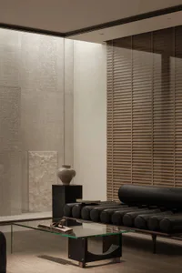 Modern minimalist living room interior with textured plaster walls, a black cylindrical leather sofa, glass coffee table, and a ceramic vase on a dark pedestal.