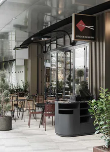 Modern sushi restaurant interior with a curved grey marble service counter, wooden slat chairs, potted trees, and a hanging sign reading 'STICKS IN SUSHI' with a red diamond logo.