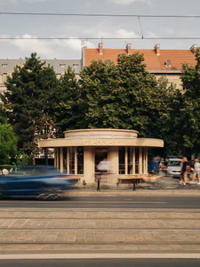 Street view of a modern cylindrical wooden bakery pavilion named Chleboteka with large glass windows, set against green trees and historic buildings in Prague.