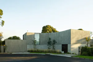 Exterior view of a modern concrete house with a long curved wall, large vertical window, wooden gate, and native landscaping at golden hour.