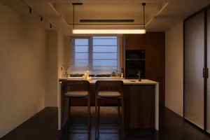 Modern minimalist kitchen featuring a walnut wood island with two bar stools, a long linear pendant light, built-in appliances, and a large window with blinds overlooking city buildings at dusk.