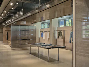 Modern industrial-style clothing boutique interior with raw concrete walls, stainless steel display table, mesh room dividers, and clothing racks under linear track lighting.