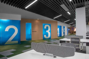Modern office interior showing a hallway with large blue meeting room doors numbered 2, 3, 4, and 5, wood paneling, green and grey patterned carpet, white desks with ergonomic chairs in the foreground, and linear ceiling lighting.