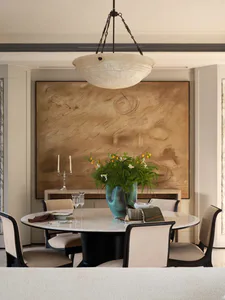 Elegant dining room featuring a round Calacatta marble table, dark wood upholstered dining chairs, a large abstract painting in warm tones, and a large turquoise ceramic vase with fresh flowers under an alabaster pendant light.