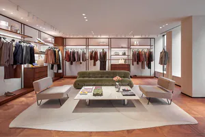 Modern luxury menswear boutique interior with olive green velvet sofa, two beige bouclé armchairs, terrazzo coffee table, herringbone wood flooring, and extensive built-in clothing display racks under warm track lighting.