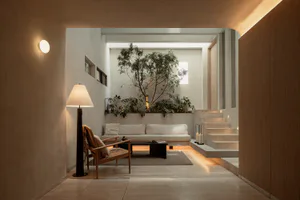 Modern minimalist living room featuring raw concrete walls, a wooden armchair, white sofa, tall floor lamp, and a large indoor tree in a raised planter, with a terrazzo staircase on the right.